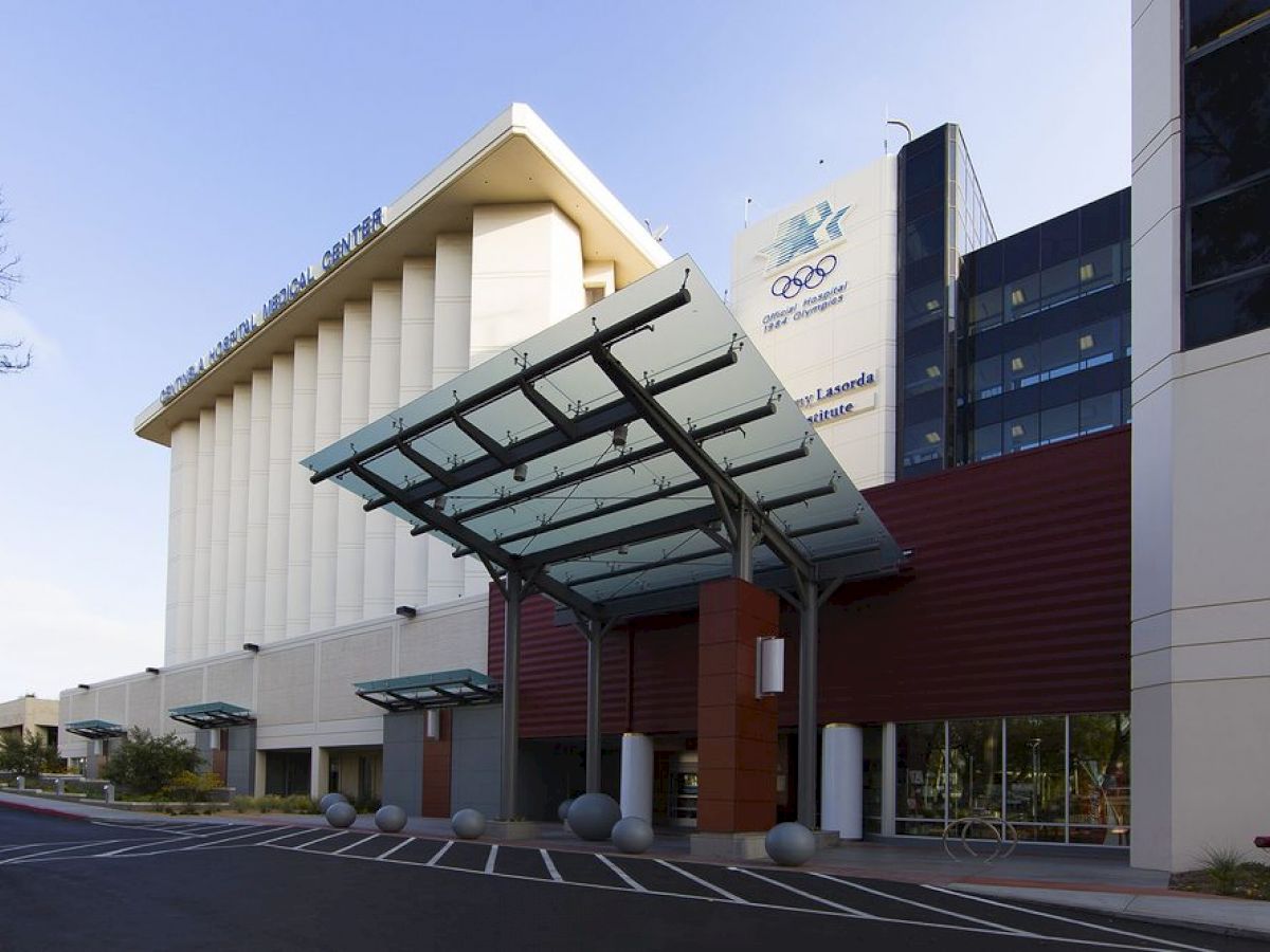 The image shows the entrance of a modern building with a glass canopy, signs with logos, and a multi-story structure in the background.