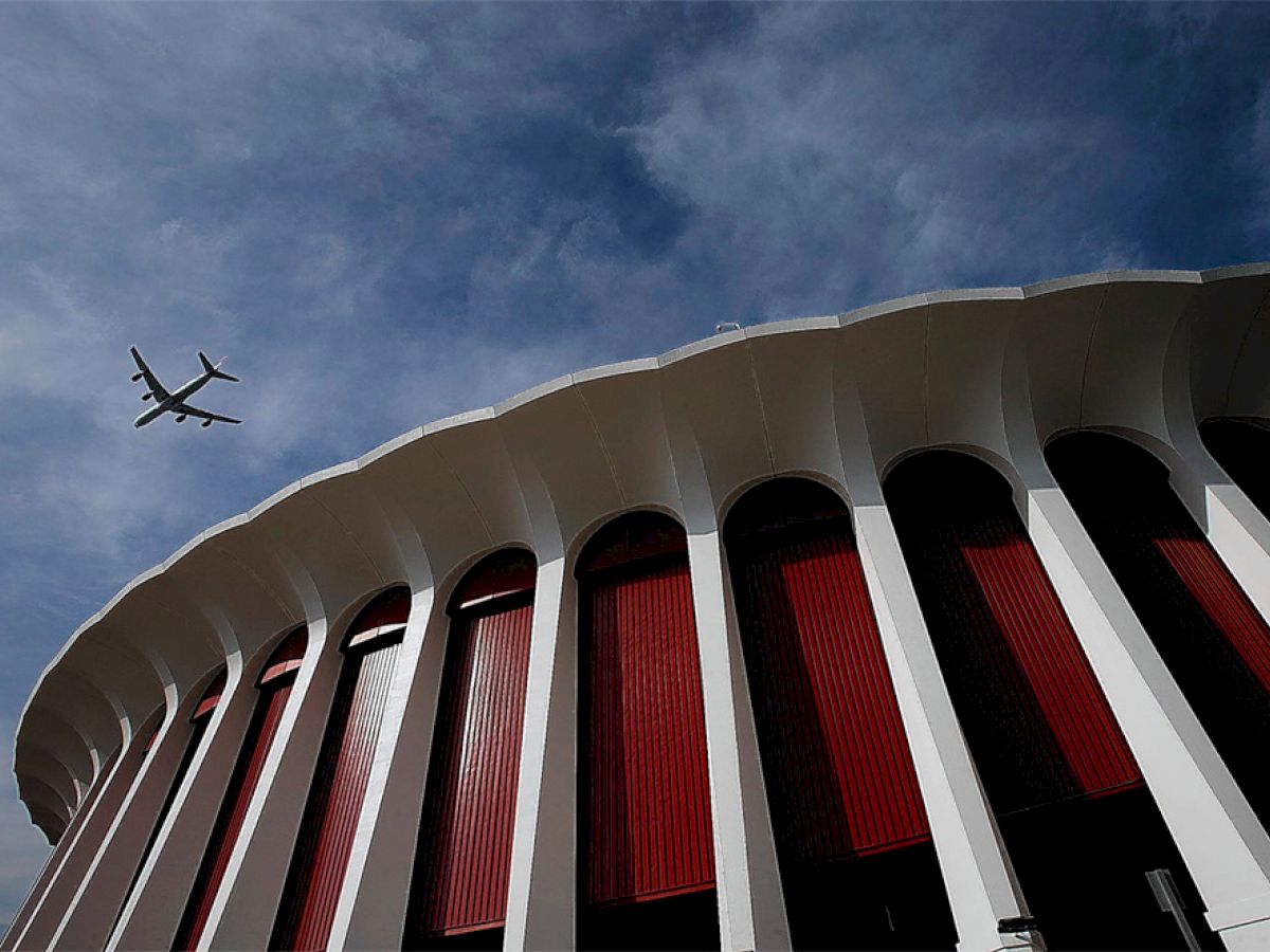 A large, circular building with tall white columns and red accents is shown from below, with an airplane flying in the sky above.