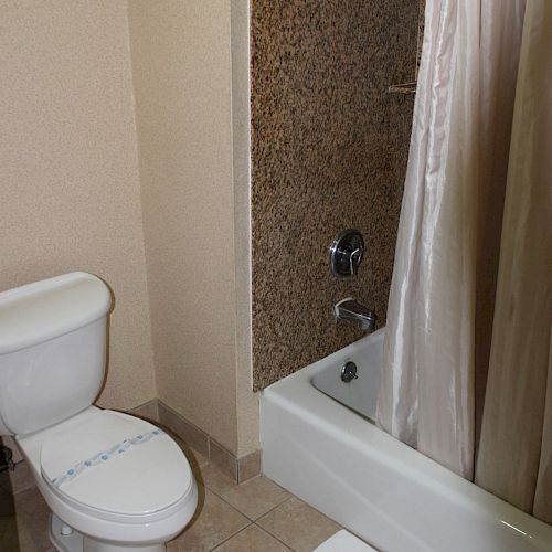 A bathroom with a toilet, a bathtub with a shower curtain, and a tiled floor and wall is shown in the image.