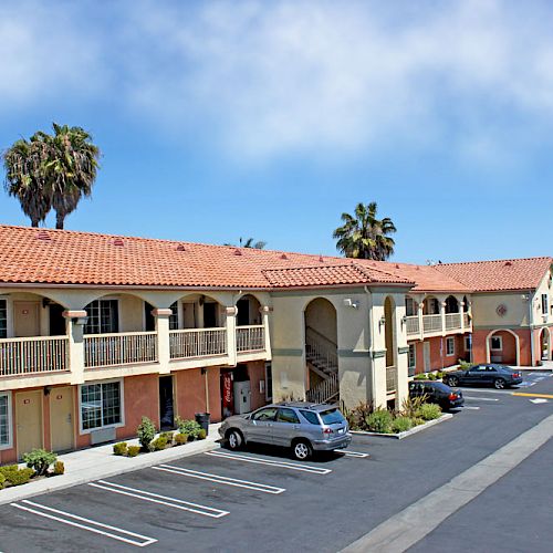 The image shows a two-story motel with a Mediterranean-style roof, ample parking, and several parked cars. Palm trees are visible in the background.