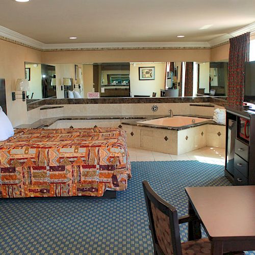 This image shows a hotel room with a bed, a large mirror, a hot tub, a TV on a dresser, and a small table with chairs.