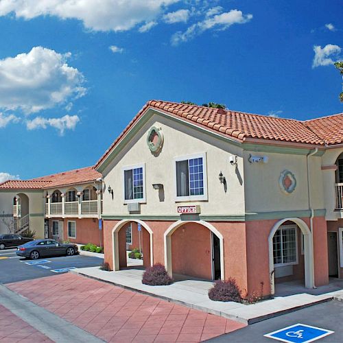 The image shows a two-story motel with a terracotta-tiled roof, arched entryways, and a parking lot, partially occupied by a few cars.