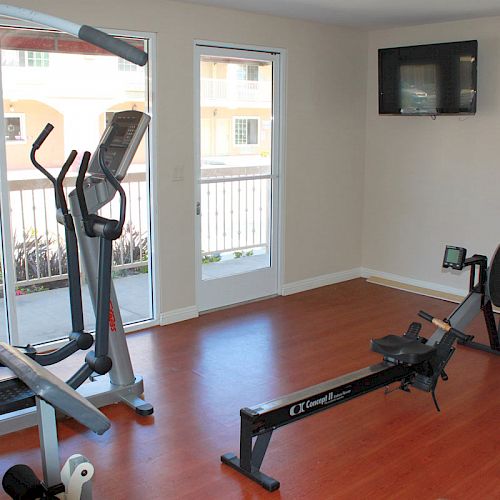 This image shows a small gym with exercise equipment including an elliptical machine and a rowing machine, with a TV mounted on the wall.