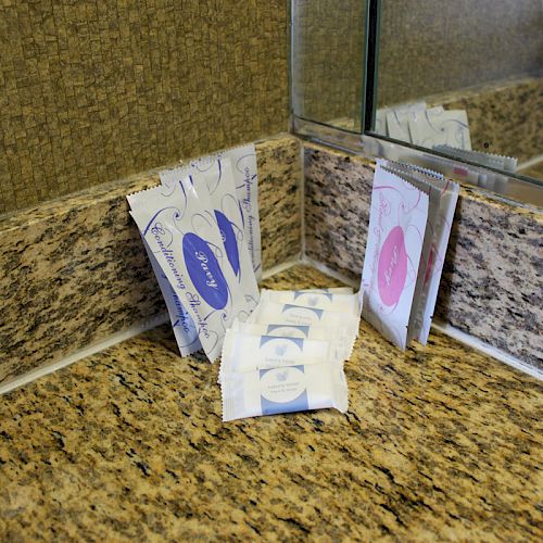 The image shows a bathroom countertop with small hygiene item packages in the corner, reflected in a mirror, next to a wall and bathroom sink.