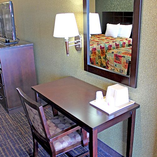 A hotel room setup with a wooden desk, cushioned chair, desk lamp, wall mirror and a folded cloth on the desk, in front of a bed.