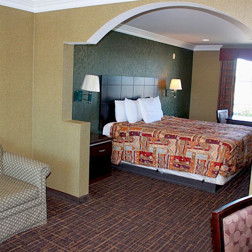 The image shows a hotel room with a bed, sofa, desk, and chairs. The bed has a patterned cover, and there is a window with curtains.