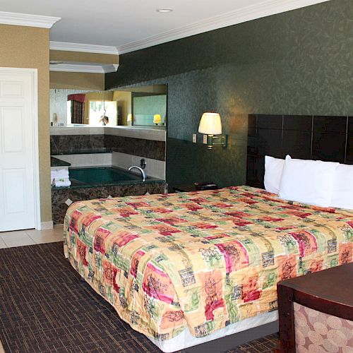 A hotel room with a king-sized bed, quilted bedspread, desk, TV, and a jacuzzi in the back corner next to a vanity mirror and sink.