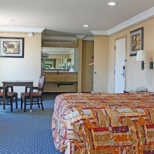 A hotel room with a large bed, two chairs at a small table, a dresser, a wall-mounted TV, and a bathroom in the background with a vanity area.