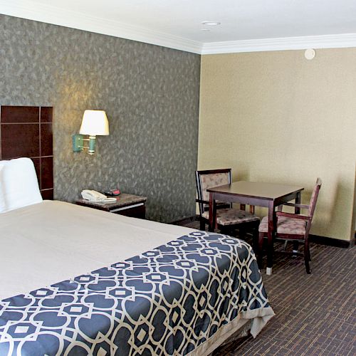 A hotel room with a bed, table with chairs, a lamp, a phone, and a coat rack in the corner. There's a patterned bedspread on the bed.