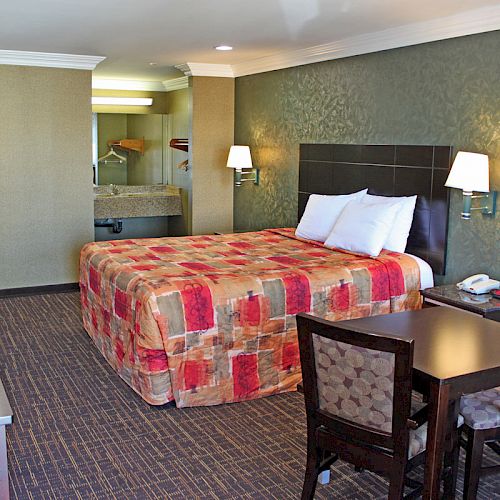 The image shows a hotel room with a bed, a desk, chairs, a TV, and a sink area in the background, all with a colorful bedspread.