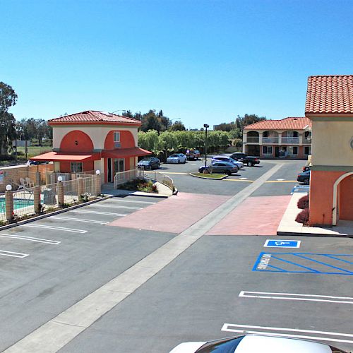 The image shows a sunny outdoor view of a motel with a parking lot, a pool area, and two buildings with red roofs and arches.