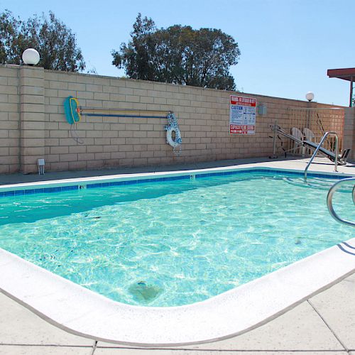 A swimming pool with clear water, a pool ladder, life preserver, and wall-mounted cleaning equipment in a fenced area with lounge chairs.