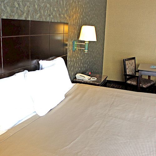 The image shows a hotel room with a bed, two pillows, a wall lamp, a table with two chairs, a telephone, and a textured wall.