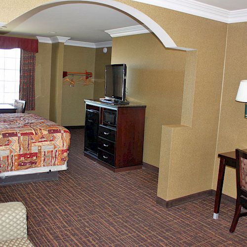 This image shows a hotel room with a bed, TV on a dresser, desk with chair, and a large window. The decor is simple and functional.