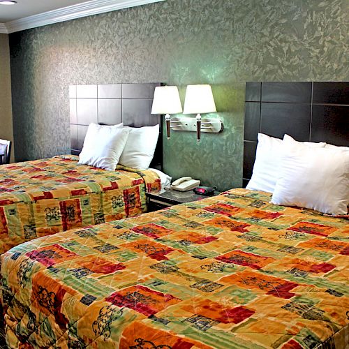 This is an image of a hotel room with two queen-sized beds, colorful bedspreads, a nightstand with a lamp, and a window with curtains.