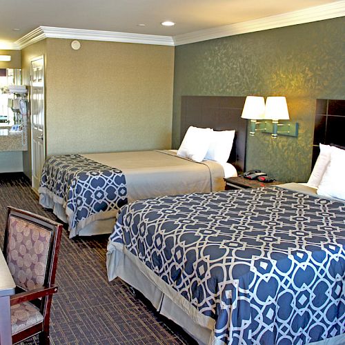 A hotel room with two double beds, patterned bedspreads, a desk, chair, TV, lamps, and an open view of a bathroom area with a sink and mirror.