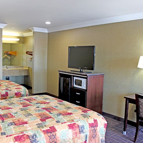 This image shows a hotel room with two double beds, a TV, a desk with a chair, a mirror, and a sink area with hanging space in the background.