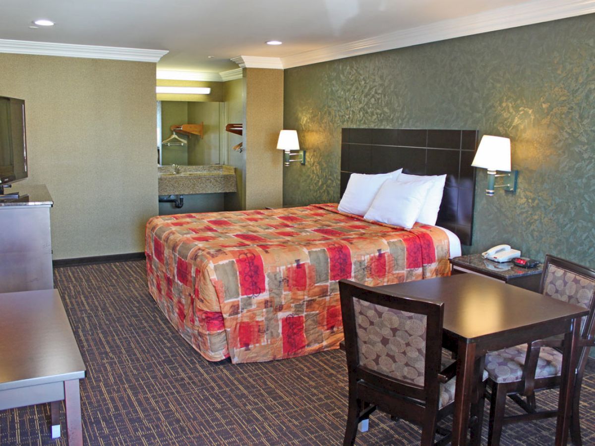 The image shows a hotel room with a bed, a TV, a small dining table with chairs, nightstands with lamps, and a bathroom in the background.