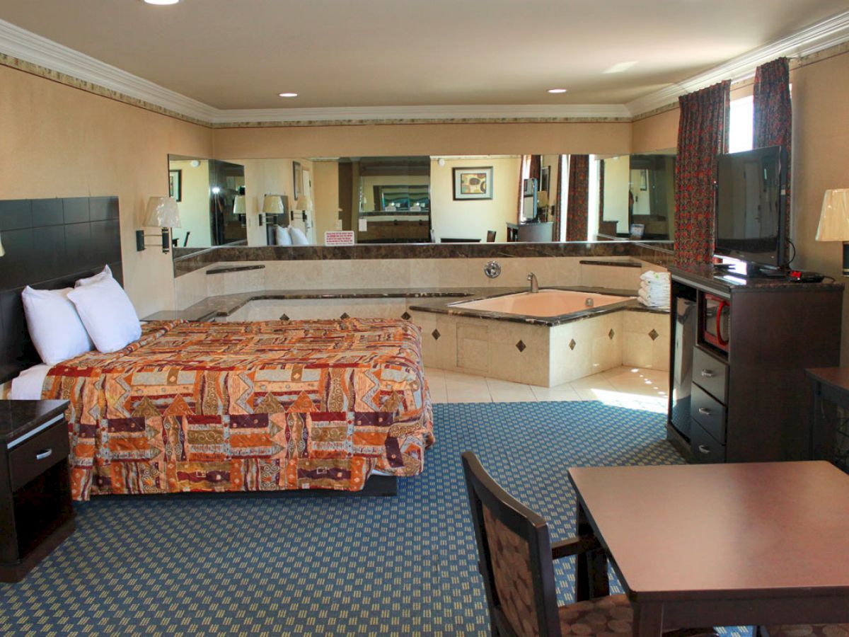 A hotel room with a bed, desk, TV, and a large jacuzzi tub. Mirrors on the walls enhance the spacious feel of the room.