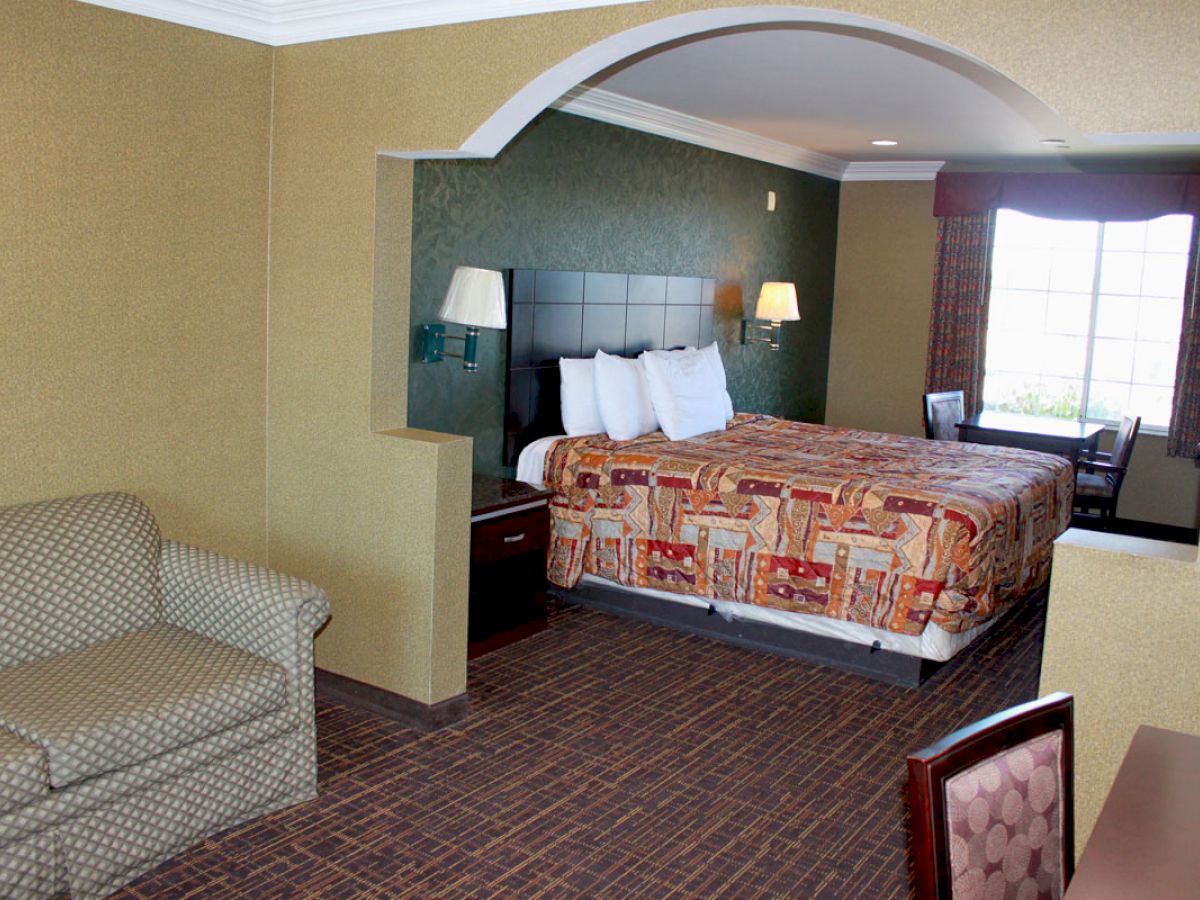 The image shows a hotel room with a bed, sofa, desk, chairs, and lamps. The bed has a patterned bedspread, and there's a window with natural light.