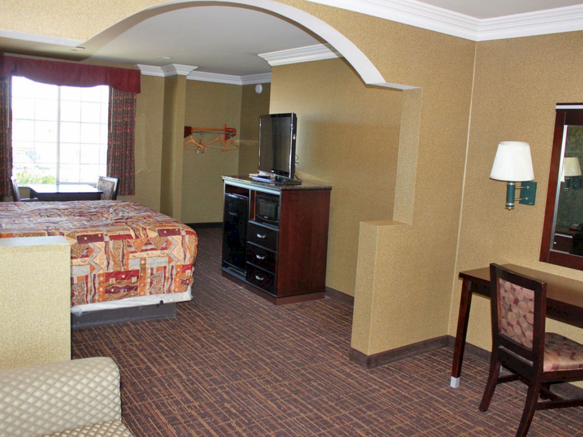 The image shows a hotel room with a bed, dresser with TV, desk with chair, mirror, lamp, and an arched entry to the bed area, overall neatly arranged.