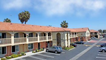 This image shows a motel with several rooms, an upper balcony, a parking lot with a few cars, and some palm trees in the background.