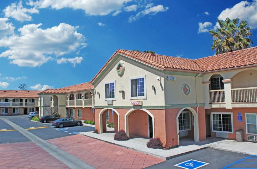 A two-story hotel with a red-tiled roof, multiple balconies, parking spaces, and clear blue skies in the background.