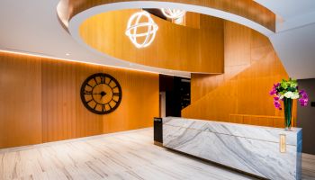 A modern lobby with a marble reception desk, wooden walls, a large clock, and a geometric light fixture hanging from a curved ceiling installation.
