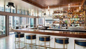 A modern, upscale bar with a wide variety of bottles on shelves, elegant stools, pendant lights, and large windows offering city views.