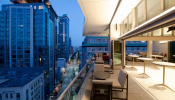 A modern cityscape with tall buildings at dusk, featuring a terrace with outdoor seating and tables, overlooking a street below.