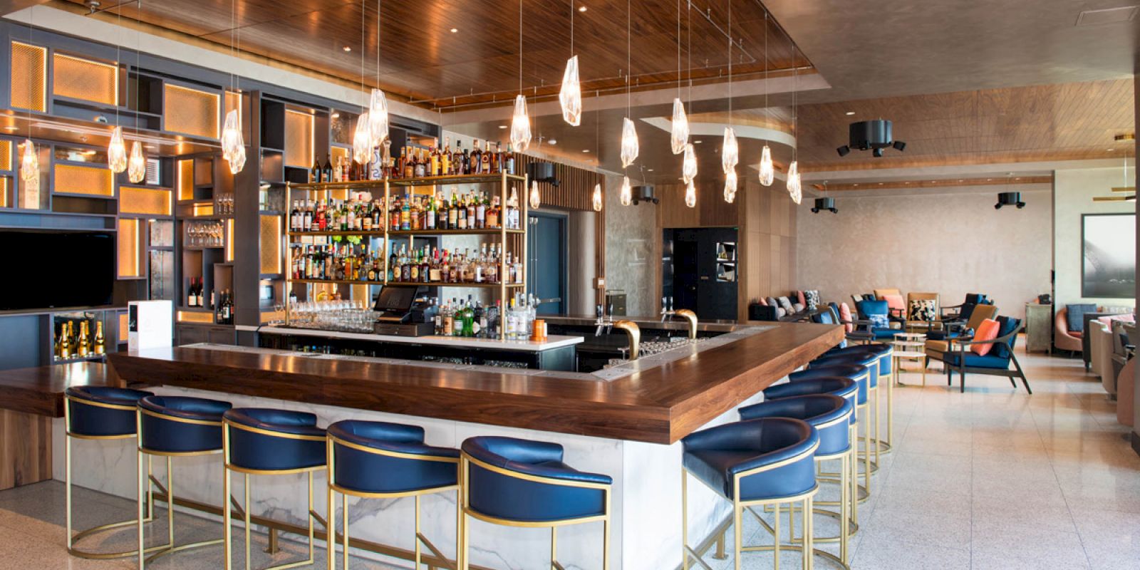 A modern bar with blue barstools, hanging lights, and an array of bottles on shelves behind the counter. Also visible are lounge areas and tables.