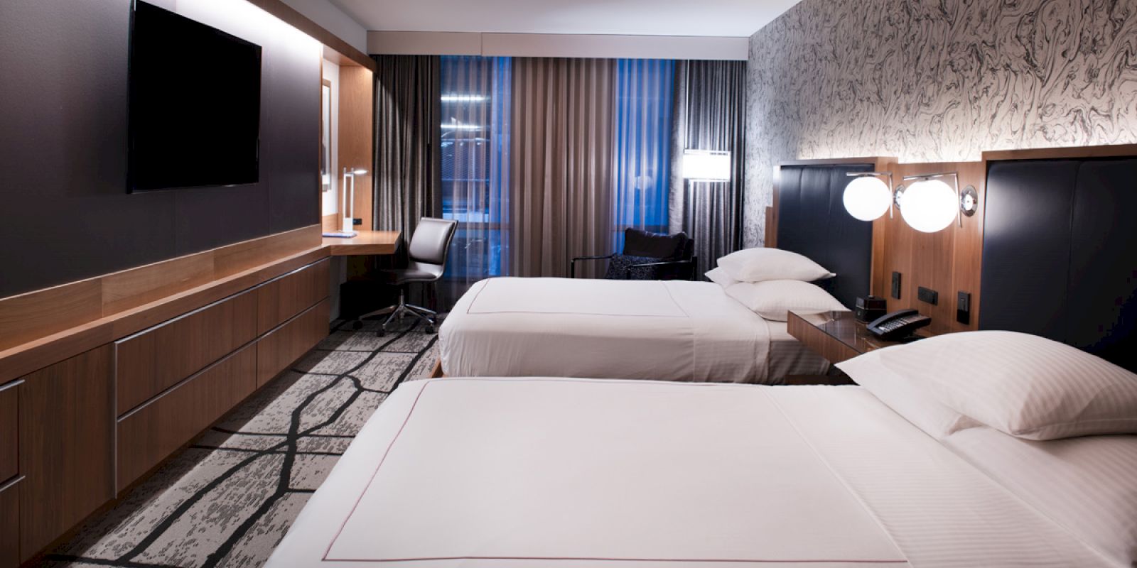 A modern hotel room with two neatly made beds, a flat-screen TV, a desk, and a chair, featuring contemporary lighting and decor on the walls.