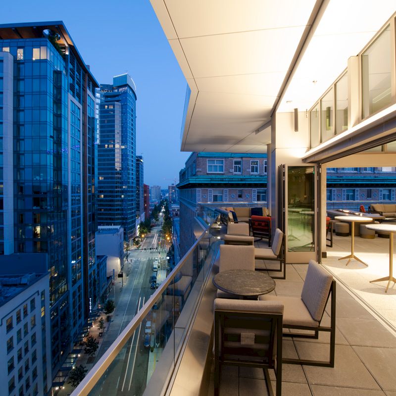 A modern cityscape with high-rise buildings, seen from a balcony at dusk. The balcony has tables, chairs, and is brightly lit by the building.