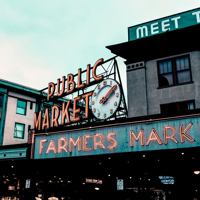 The image shows a historic market with neon signs reading "PUBLIC MARKET" and "FARMERS MARKET" on building exteriors under a cloudy sky.