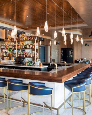 The image shows a modern bar with high-back chairs, a fully stocked counter, and contemporary overhead lighting fixtures in an elegant setting.