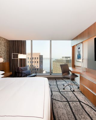 A modern hotel room with a large bed, wall-mounted TV, desk, and chair overlooking buildings through a large window, featuring contemporary decor.