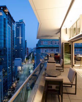 A rooftop terrace overlooking a modern cityscape at dusk, with outdoor seating areas and tall buildings in the background, under a twilight sky.