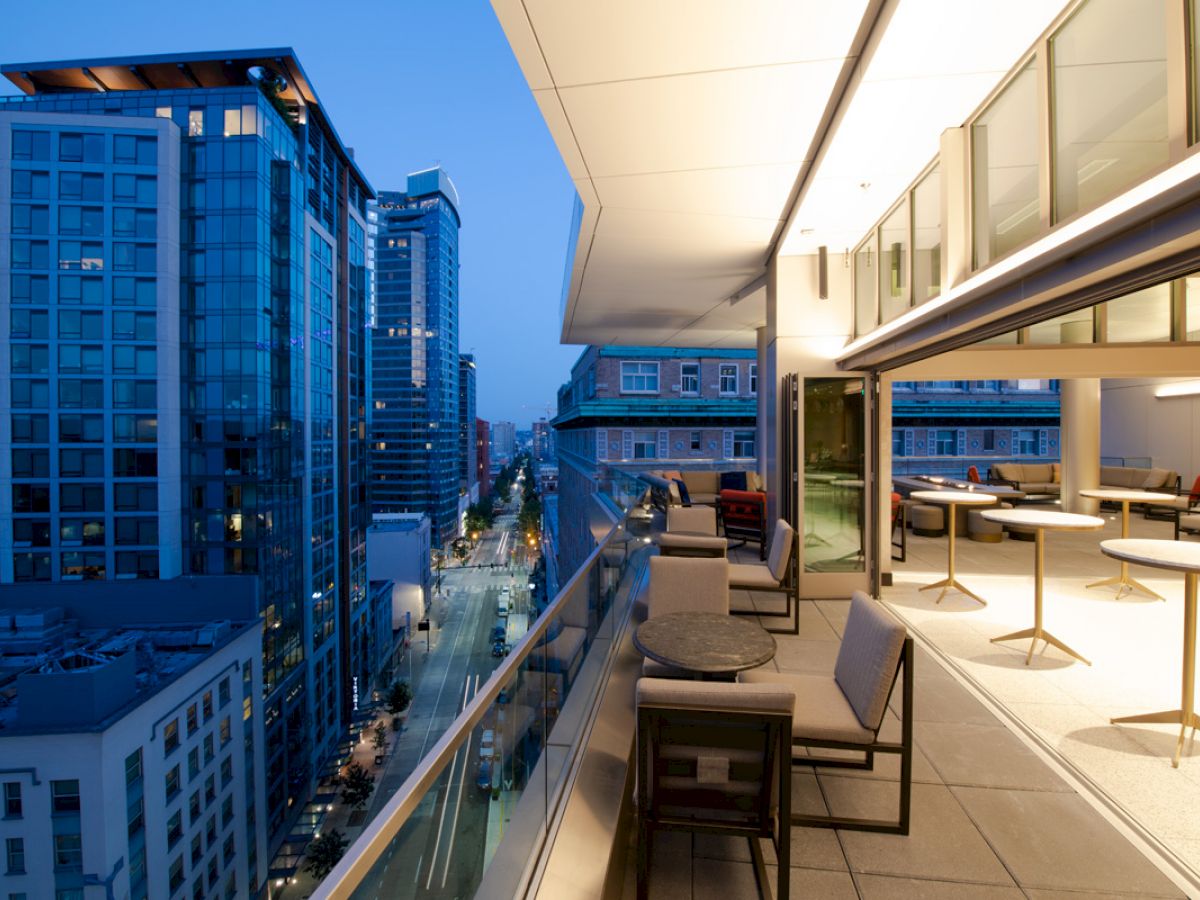 A modern terrace overlooking a city street, with buildings illuminated at dusk. The terrace features tables, chairs, and a sleek design.
