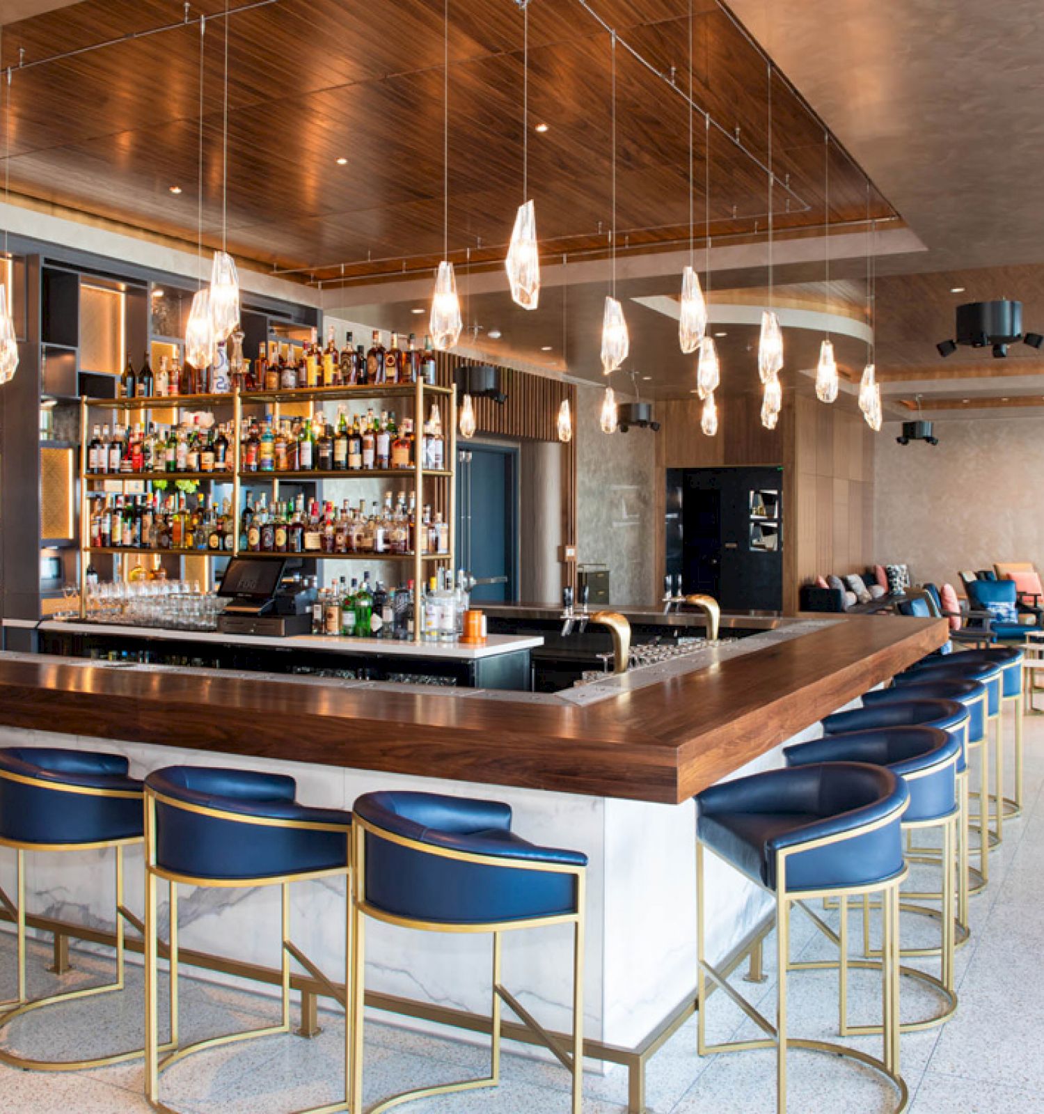 This image shows a modern bar with a large counter, blue bar stools, various hanging lights, and a fully stocked shelf of liquor bottles in the back.