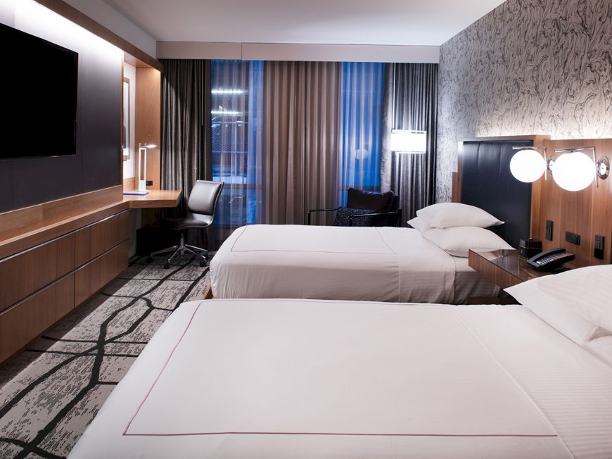 A modern hotel room with two beds, a wall-mounted TV, a desk with a chair, and large windows with curtains.
