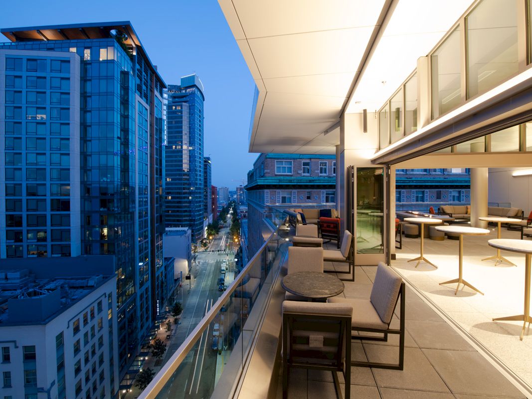 A modern urban balcony with outdoor seating overlooks a city street lined with tall buildings in the evening.