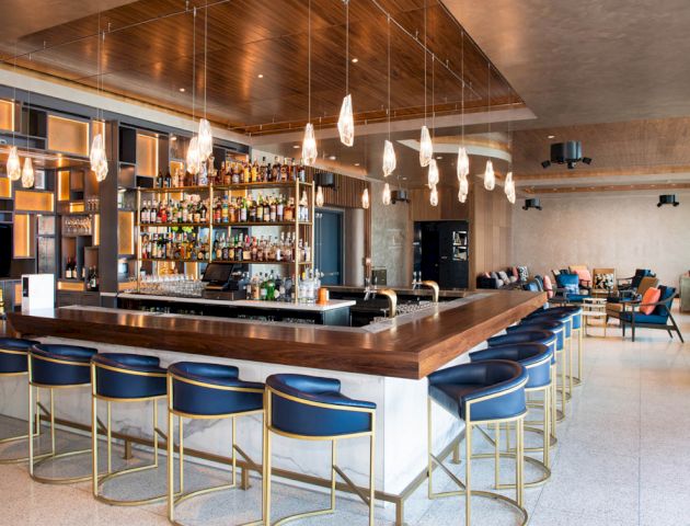 A modern bar with a wooden countertop, shelves stocked with bottles, blue-cushioned bar stools, and stylish hanging lights in a spacious setting.