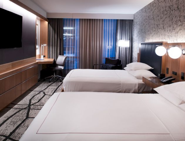 A modern hotel room with two beds, wall-mounted TV, desk, and chair by the window; patterned carpet and wallpaper add to the sophisticated decor.