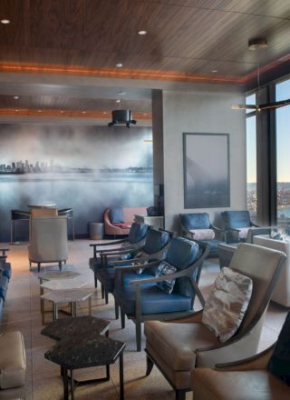 A modern lounge with comfortable seating, wall art, and large windows offering views of a city skyline and waterfront Ferris wheel, at sunset.