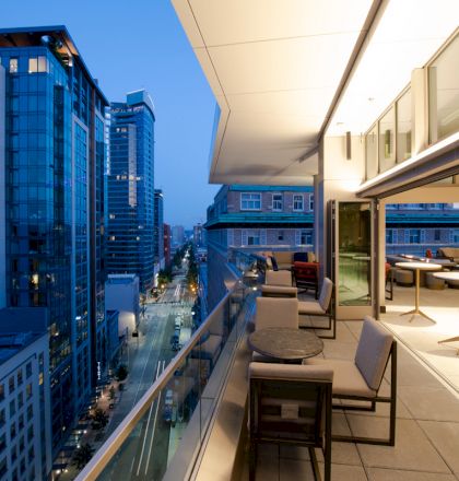 A modern balcony view overlooking a city street at dusk, with tables and chairs. Tall buildings and a lit interior are visible.
