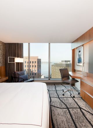 A modern hotel room with a king-sized bed, a large TV, a desk, and a view of the city through the window, featuring contemporary decor.