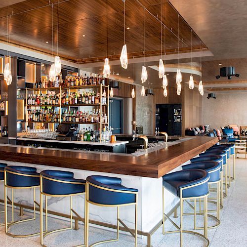 A modern bar with blue padded stools, wooden countertops, hanging pendant lights, and a well-stocked shelf of liquor bottles in a stylish lounge area.
