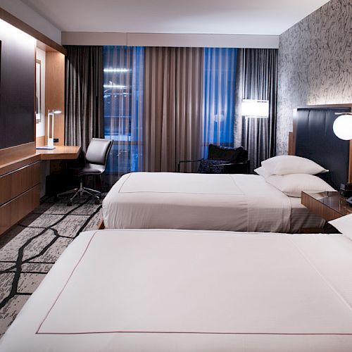 A modern hotel room with two beds, a wall-mounted TV, a work desk, a stylish chair, and contemporary decor and lighting fixtures.