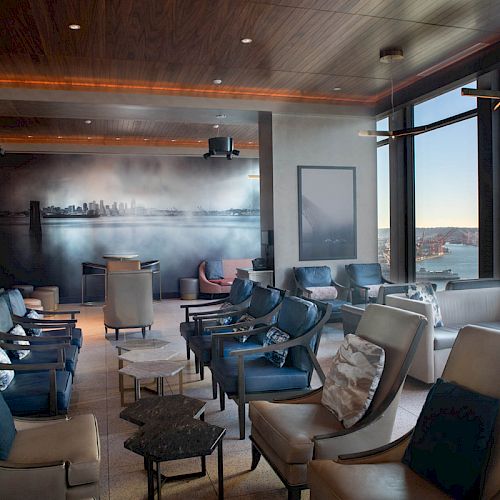 A modern lounge with comfortable seating, large windows offering a scenic water view, and a wall mural depicts a city skyline in the background.