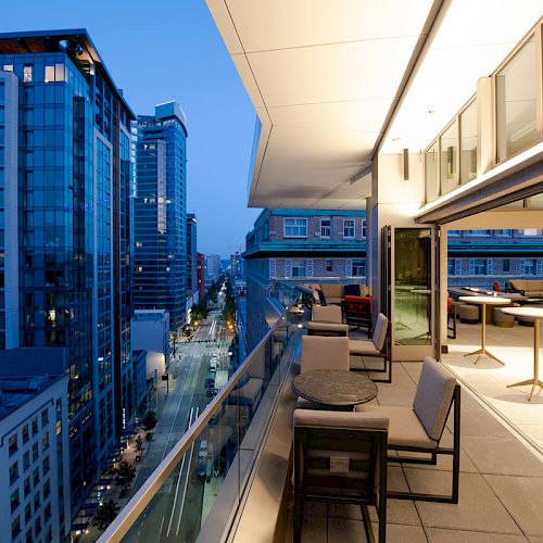 A modern cityscape at dusk with high-rise buildings, a well-lit balcony with seating, tables, and a glass railing overlooking the street.